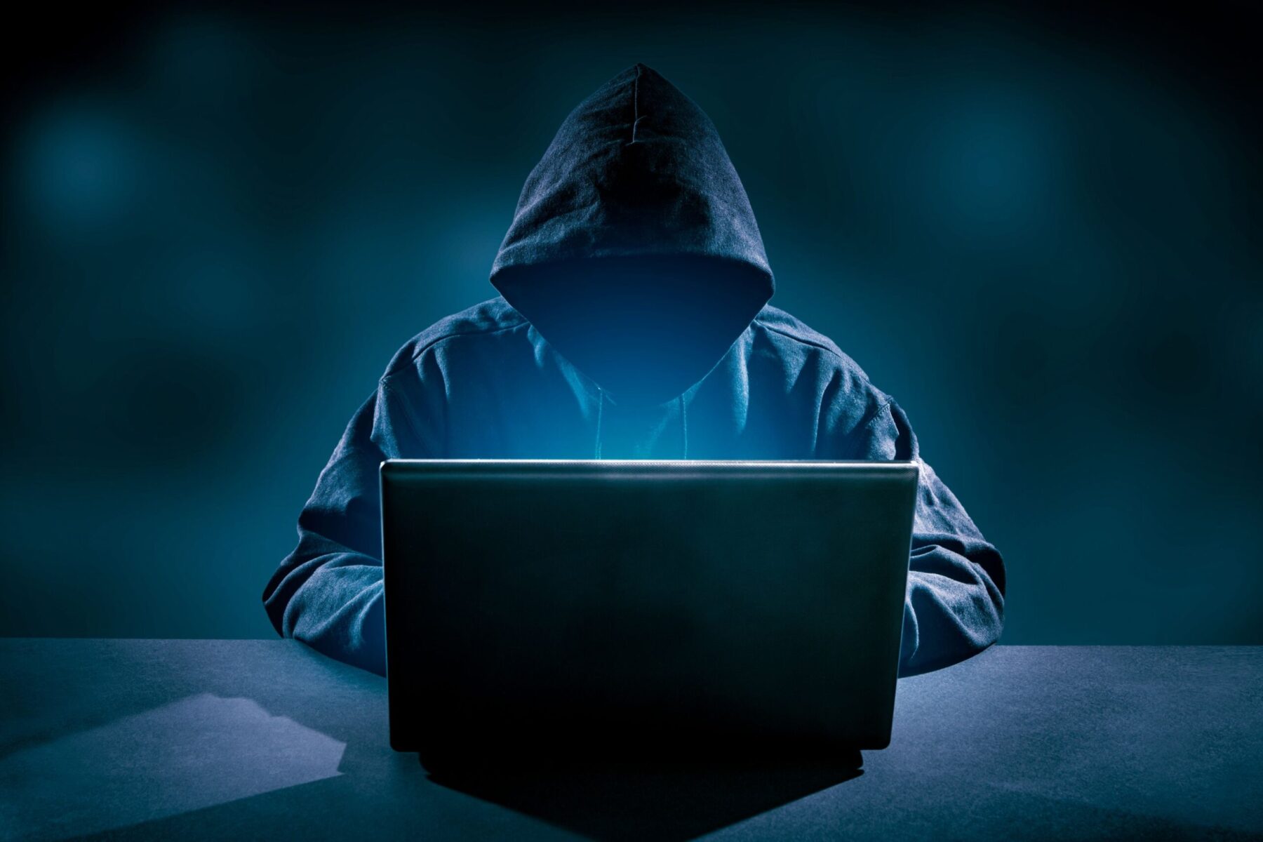 man in a dark hoodie with his face hidden by shadows sits in front of a laptop