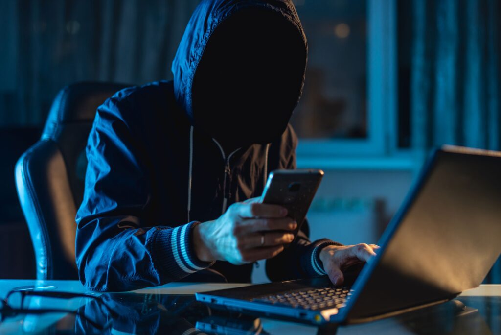 Man in a hoodie with his face hidden by shadows holds a cellphone while sitting in front of a laptop to steal someones identity